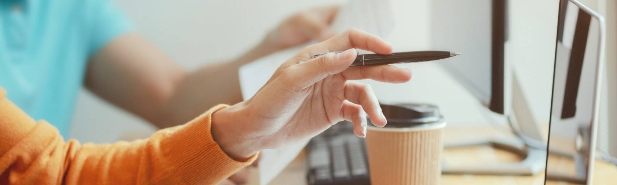 Close up of woman's hand wearing orange jumper holding pen pointing at laptop